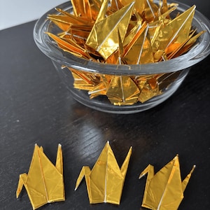 May include: A clear glass bowl filled with numerous gold origami cranes. Three additional gold cranes are arranged on a dark surface. The cranes are folded with sharp angles and white accents, creating a striking visual contrast.
