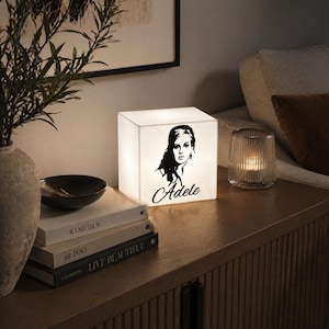 May include: A white cube-shaped lamp featuring a black silhouette of Adele and her name in script. The lamp is illuminated, sitting on a wooden cabinet next to books and a candle in a glass holder. The room has a neutral color palette.