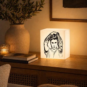 May include: A white cube lamp featuring a black and white illustration of a person with tattoos. The lamp is illuminated, casting a soft glow. It sits on a wooden surface with a vase, books, and a candle.