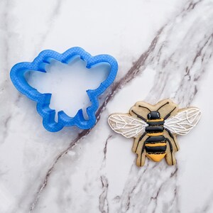 May include: A blue plastic cookie cutter shaped like a bee, next to a decorated sugar cookie shaped like a bee with black and yellow stripes and white wings.
