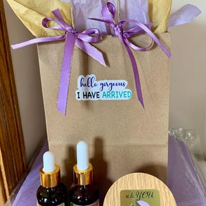 Peut inclure: Un sac cadeau en papier brun avec un ruban violet et du papier de soie. Le sac porte un autocollant avec l'inscription "hello gorgeous I HAVE ARRIVED". Deux petites bouteilles marron avec des bouchons dor&eacute;s et un pot en bois sont devant le sac. Les bouteilles et le pot portent le logo "u.b. YOU".