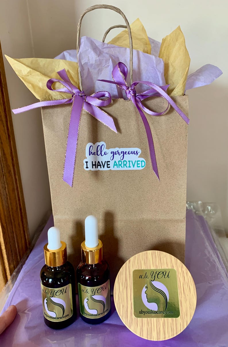 Peut inclure: Un sac cadeau en papier brun avec un ruban violet et du papier de soie. Le sac porte un autocollant avec l'inscription "hello gorgeous I HAVE ARRIVED". Deux petites bouteilles marron avec des bouchons dor&eacute;s et un pot en bois sont devant le sac. Les bouteilles et le pot portent le logo "u.b. YOU".