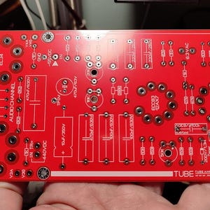 Pode incluir: Uma placa de circuito impresso vermelha com vários componentes eletrônicos e texto. A placa apresenta numerosos orifícios, conectores e etiquetas como "KT88 EL34" e "AUDIO CHANNEL". A placa é retangular e tem um design complexo.