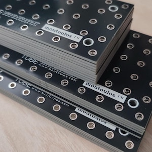 以下が含まれることがあります： 複数の穴が均等に配置された黒い金属板で、「moutoulos」と「TUBE」の文字が印刷されています。プレートは積み重ねられています。プレートは明るい木材の表面にあります。