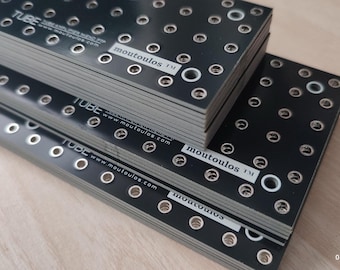 May include: Black metal plates with multiple evenly spaced holes and the text "moutoulos" and "TUBE" printed on them. The plates are stacked on top of each other. The plates are on a light wood surface.