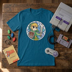 Op de afbeelding: Tealkleurig t-shirt met een cirkelvormige afbeelding van een stripfiguur met een zwaard en schild. Een Super Nintendo-console, controller en gamecartridge liggen naast het shirt. Een boekje en sleutels liggen ook op het houten oppervlak.