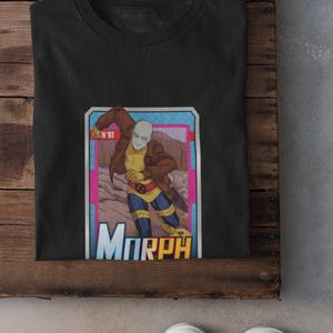 May include: A black t-shirt featuring a graphic print of the character Morph from X-Men '97. The design shows the character in a brown jacket, yellow shirt, and blue pants, framed within a rectangular border with the text Morph at the bottom.