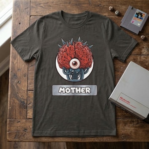 Puede incluir: Camiseta gris oscuro con un gráfico de un cerebro rojo con un ojo, púas y elementos robóticos. La palabra "MOTHER" está impresa debajo. Se ven un cartucho y una consola de juegos de Nintendo.