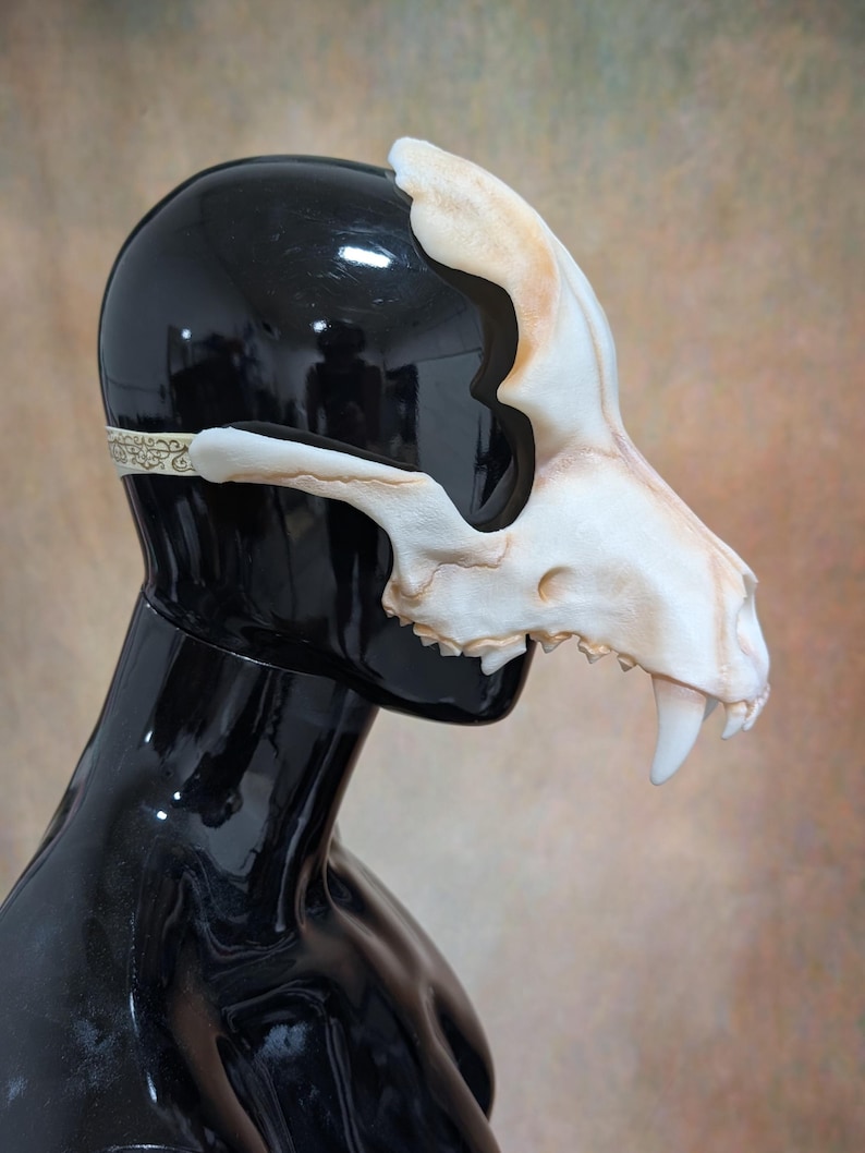 May include: A bone-colored, animal skull mask with visible teeth and a detailed design. The mask is worn on a black mannequin head, with a decorative band around the back. The mask has a realistic, textured appearance.