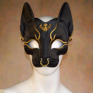 K&ouml;nnte beinhalten: Schwarze Katzenmaske mit goldenen Akzenten und einem Skarab&auml;us-Design. Die Maske hat gro&szlig;e, spitze Ohren, goldfarbene Reifen und einen Nasenring. Die Augen&ouml;ffnungen sind wei&szlig;. Die Maske wird auf einem wei&szlig;en Schaufensterpuppenkopf pr&auml;sentiert.
