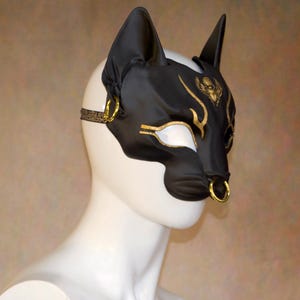 K&ouml;nnte beinhalten: Schwarze Katzenohren-Maske mit goldenen Akzenten, darunter ein stilisierter Sch&auml;del und Augendetails. Die Maske hat einen Nasenring und einen gemusterten Riemen. Die Maske wird auf einem wei&szlig;en Schaufensterkopf pr&auml;sentiert.