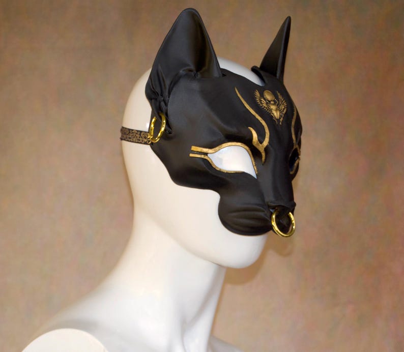 K&ouml;nnte beinhalten: Schwarze Katzenohren-Maske mit goldenen Akzenten, darunter ein stilisierter Sch&auml;del und Augendetails. Die Maske hat einen Nasenring und einen gemusterten Riemen. Die Maske wird auf einem wei&szlig;en Schaufensterkopf pr&auml;sentiert.