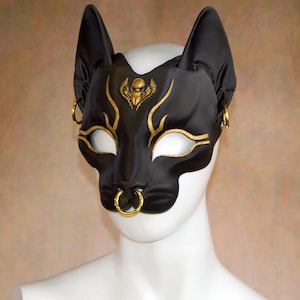 K&ouml;nnte beinhalten: Schwarze Katzenmaske mit goldenen Akzenten, einschlie&szlig;lich eines Skarab&auml;us-Emblems und dekorativer Linien. Die Maske hat offene Augenl&ouml;cher und einen Nasenring, mit ohrartigen Verl&auml;ngerungen. Die Maske wird auf einem wei&szlig;en Schaufensterpuppenkopf pr&auml;sentiert.