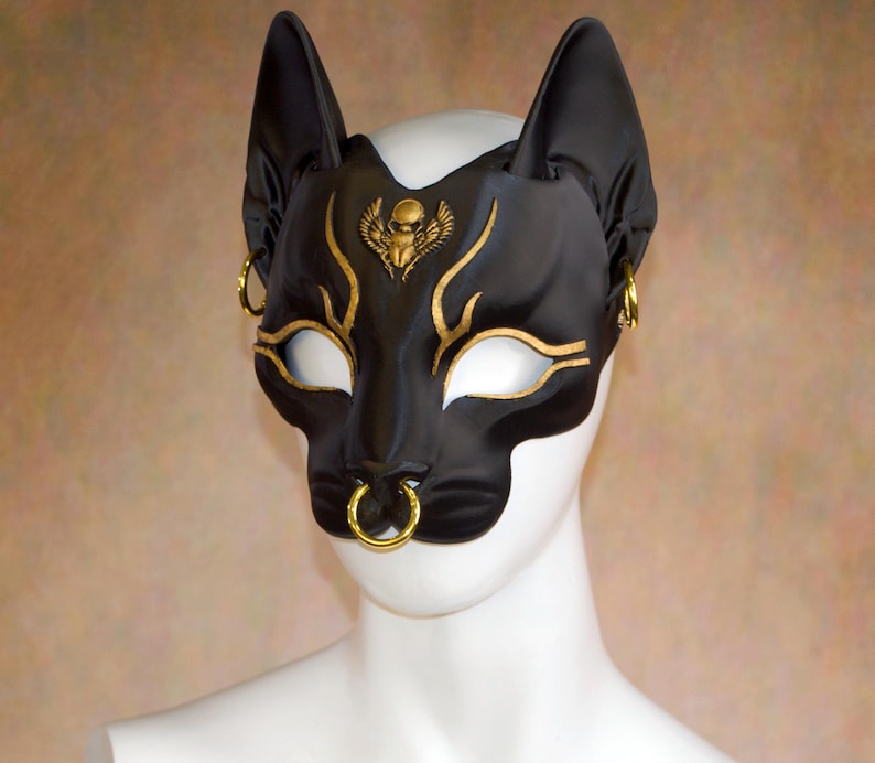 K&ouml;nnte beinhalten: Schwarze Katzenmaske mit goldenen Akzenten, einschlie&szlig;lich eines Skarab&auml;us-Emblems und dekorativer Linien. Die Maske hat offene Augenl&ouml;cher und einen Nasenring, mit ohrartigen Verl&auml;ngerungen. Die Maske wird auf einem wei&szlig;en Schaufensterpuppenkopf pr&auml;sentiert.