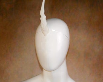 May include: A white, spiraled, tapered horn accessory attached to a thin, matching white headband. The horn features a textured, twisted surface that rises vertically from the center of the headband, creating a distinct, singular point.