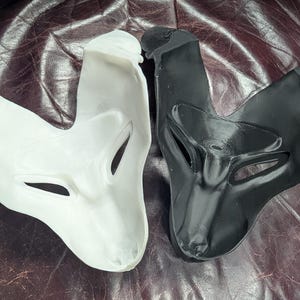 May include: Two fox-shaped masks, one white and one black, are displayed. The masks have eye and nose openings and are made of a smooth material. The masks are likely for costume or theatrical use.