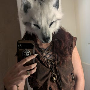 May include: A white and gray fox mask with closed eyes. The mask is worn with a brown vest and a decorative necklace. A brown phone case with a lion emblem is also visible.