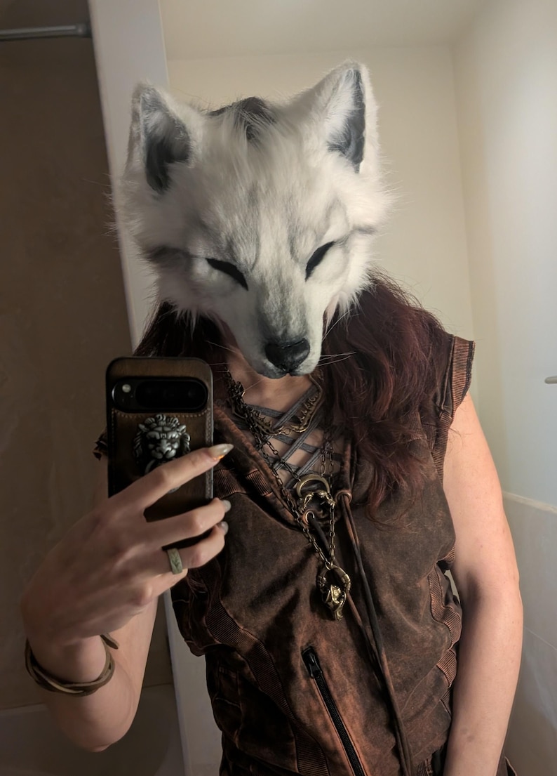 May include: A white and gray fox mask with closed eyes. The mask is worn with a brown vest and a decorative necklace. A brown phone case with a lion emblem is also visible.