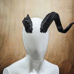 Könnte beinhalten: Ein Paar schwarze, strukturierte, gebogene Hörner aus Kunststoff. Die Hörner sind an einem Stirnband befestigt und werden auf dem Kopf getragen.