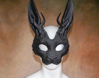 May include: A black mask with rabbit ears and small antlers. The mask has white eye openings and is designed to cover the upper face. The mask is made of a textured material and is displayed on a white mannequin head.