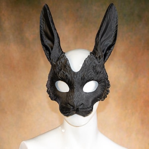 Könnte beinhalten: Eine schwarze Hasenmaske mit langen, aufrechten Ohren und einem strukturierten, fellartigen Aussehen. Die Maske hat große Augenöffnungen und wird auf einem weißen Schaufensterpuppenkopf präsentiert. Die Maske ist wahrscheinlich für Kostüm- oder Cosplay-Zwecke.
