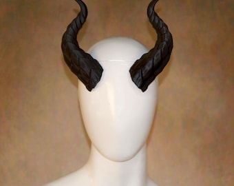 May include: A pair of matte black, curved, textured horns with a segmented, ridged surface design. The horns are positioned symmetrically on the sides of a smooth, featureless white mannequin head, extending upwards and slightly outward in a curve.