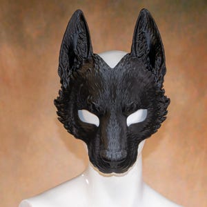 May include: A black wolf mask with detailed fur texture and open eye sockets. The mask is designed to cover the upper face, featuring pointed ears and a realistic snout. The mask is displayed on a white mannequin head against a neutral background.