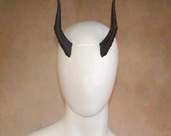 May include: A pair of black, textured, curved horns attached to a thin, clear headband. The horns feature a detailed, ridged surface that tapers to a point at the top. The headband is positioned horizontally across the top of a smooth, white mannequin head.