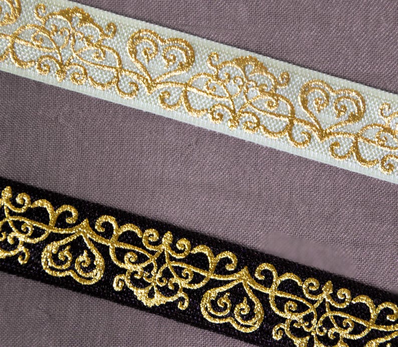 K&ouml;nnte beinhalten: Zwei dekorative B&auml;nder mit goldgestickten Designs. Ein Band ist wei&szlig;, das andere ist schwarz. Die goldene Stickerei zeigt ein sich wiederholendes Muster aus Herzen und wirbelnden Linien, das einen eleganten und verzierten Look erzeugt.