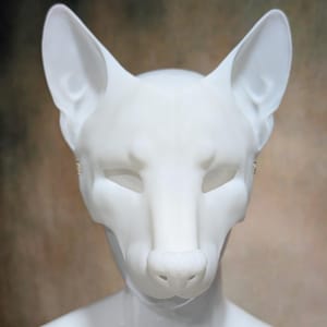 May include: A white fox mask with pointed ears, a prominent nose, and closed eye sockets. The mask is mounted on a white mannequin head, set against a blurred brown and beige background. The mask is a costume or cosplay item.