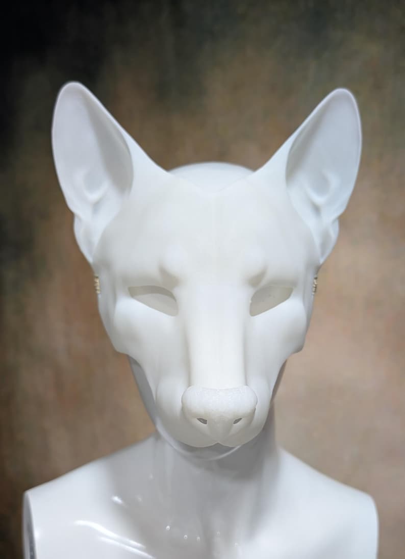 May include: A white fox mask with pointed ears, a prominent nose, and closed eye sockets. The mask is mounted on a white mannequin head, set against a blurred brown and beige background. The mask is a costume or cosplay item.