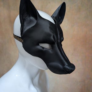 May include: A black and white dog mask with pointed ears and a long snout. The mask is made of a shiny material and has eye openings. A thin strap with gold detailing secures the mask.