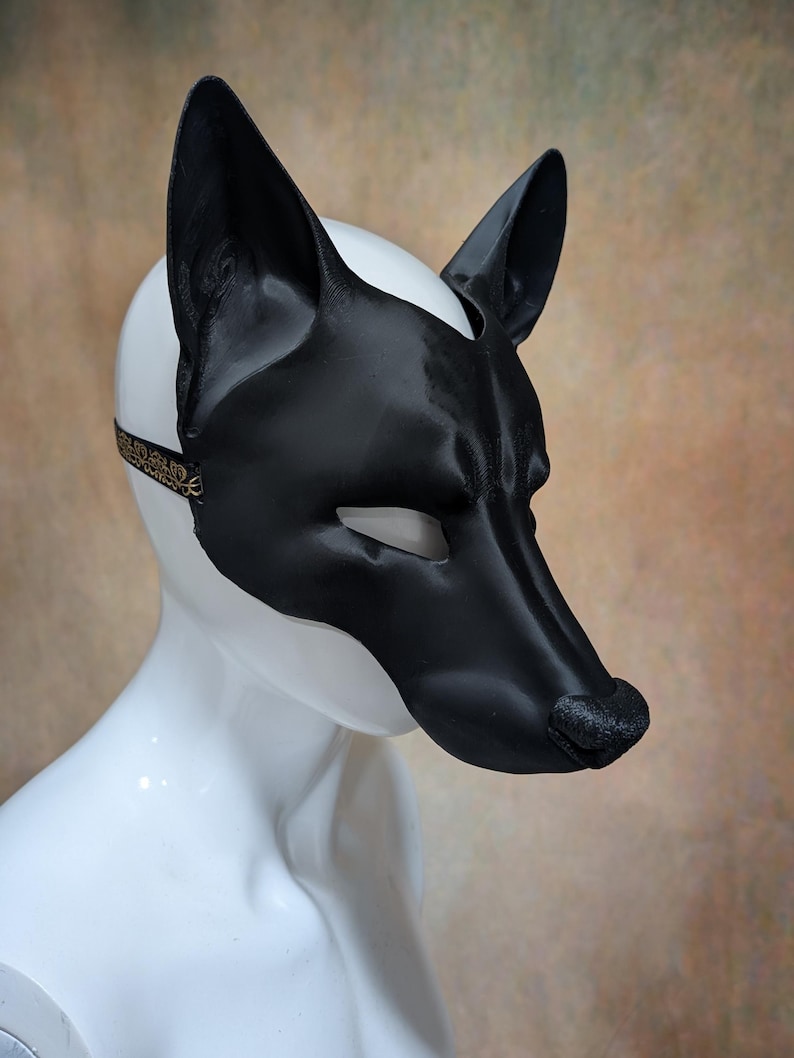 May include: A black and white dog mask with pointed ears and a long snout. The mask is made of a shiny material and has eye openings. A thin strap with gold detailing secures the mask.