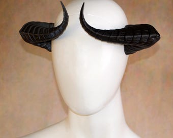 May include: A pair of black, curved, ridged horns attached to a thin headband. The horns feature a segmented, textured surface that tapers to a point, extending outward from the sides of a smooth, featureless white mannequin head display.