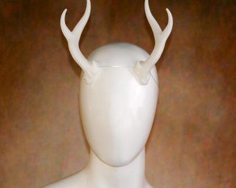 May include: A white headband featuring two symmetrical, upward-curving deer antlers attached to the top. The antlers are smooth, off-white, and branch into two points each. The headband is displayed on a smooth, featureless white mannequin head.