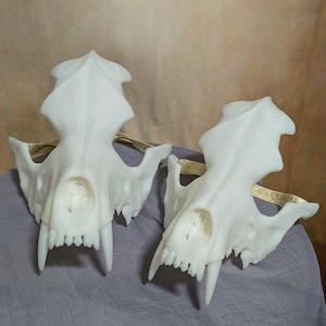 May include: Two white, stylized skull masks with sharp teeth and pointed ears. The masks are connected by a gold-colored band. The skulls are resting on a gray surface.