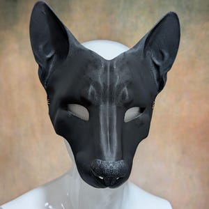 May include: A black leather mask in the shape of a canine's face, featuring pointed ears, eye openings, and a textured nose. The mask is displayed on a white mannequin head against a blurred, neutral-toned background.