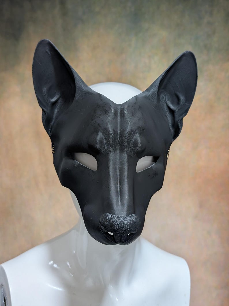 May include: A black leather mask in the shape of a canine's face, featuring pointed ears, eye openings, and a textured nose. The mask is displayed on a white mannequin head against a blurred, neutral-toned background.
