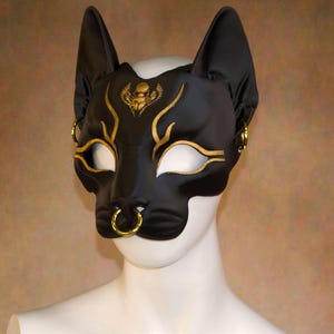 K&ouml;nnte beinhalten: Schwarze Katzenmaske mit goldenen Akzenten. Die Maske hat ein katzenartiges Design mit spitzen Ohren und einem Nasenring. Goldene Details umfassen Augenlinien, Stirndesign und Ohrringe. Die Maske besteht aus einem glatten, gl&auml;nzenden Material.