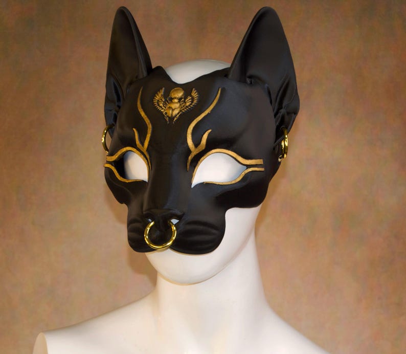 K&ouml;nnte beinhalten: Schwarze Katzenmaske mit goldenen Akzenten. Die Maske hat ein katzenartiges Design mit spitzen Ohren und einem Nasenring. Goldene Details umfassen Augenlinien, Stirndesign und Ohrringe. Die Maske besteht aus einem glatten, gl&auml;nzenden Material.