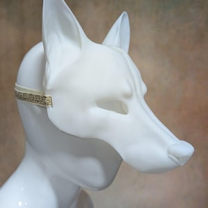 May include: A white, stylized fox mask with pointed ears and a long snout. The mask is secured with a thin, patterned elastic band. The mask is made of a smooth, matte material.