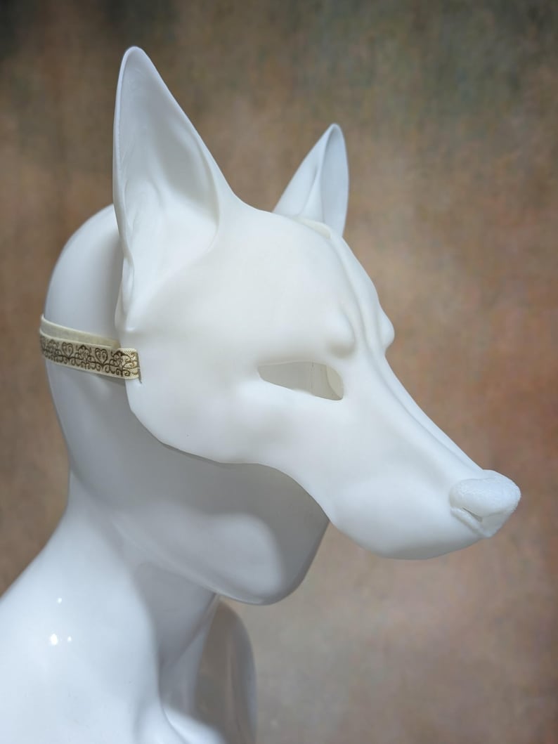 May include: A white, stylized fox mask with pointed ears and a long snout. The mask is secured with a thin, patterned elastic band. The mask is made of a smooth, matte material.