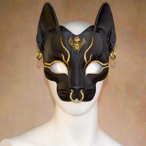 K&ouml;nnte beinhalten: Schwarze Katzenmaske mit goldenen Akzenten und Creolen. Die Maske hat ein goldenes Sch&auml;del- und Fl&uuml;geldesign &uuml;ber den Augen. Die Maske hat einen Nasenring und wird auf einem wei&szlig;en Schaufensterkopf getragen. Die Maske besteht aus gl&auml;nzendem Material.