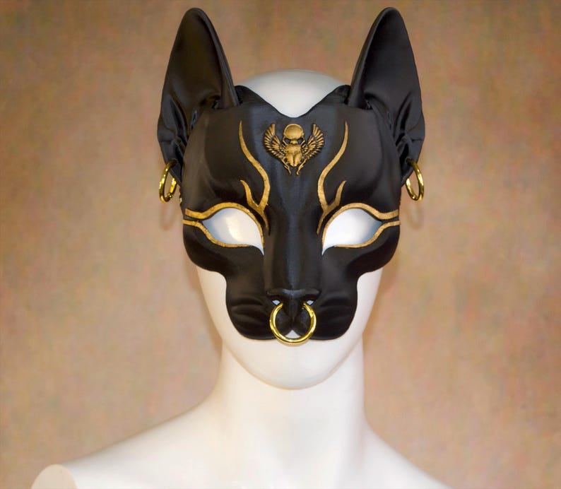 K&ouml;nnte beinhalten: Schwarze Katzenmaske mit goldenen Akzenten und Creolen. Die Maske hat ein goldenes Sch&auml;del- und Fl&uuml;geldesign &uuml;ber den Augen. Die Maske hat einen Nasenring und wird auf einem wei&szlig;en Schaufensterkopf getragen. Die Maske besteht aus gl&auml;nzendem Material.