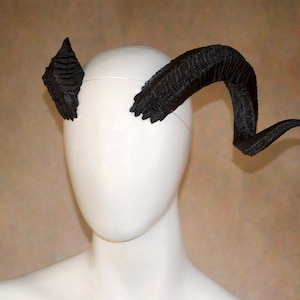 Peut inclure: Une paire de cornes noires, texturées et incurvées fixées sur une tête de mannequin blanche. Les cornes présentent un motif de surface strié et superposé qui se termine en pointe. La corne gauche est plus courte et inclinée vers le haut, tandis que la corne droite est plus longue et se courbe vers l'extérieur et le bas.
