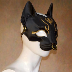 K&ouml;nnte beinhalten: Schwarze Katzenohren-Maske mit goldenen Akzenten. Die Maske hat einen goldenen Nasenring und goldene Verzierungen um die Augen und die Stirn. Die Maske besteht aus einem gl&auml;nzenden schwarzen Material und wird auf einem wei&szlig;en Schaufensterkopf pr&auml;sentiert.