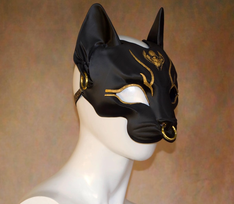 K&ouml;nnte beinhalten: Schwarze Katzenohren-Maske mit goldenen Akzenten. Die Maske hat einen goldenen Nasenring und goldene Verzierungen um die Augen und die Stirn. Die Maske besteht aus einem gl&auml;nzenden schwarzen Material und wird auf einem wei&szlig;en Schaufensterkopf pr&auml;sentiert.