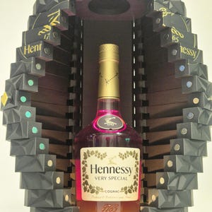 Könnte beinhalten: Eine Flasche Hennessy Very Special Cognac in einem schwarzen, geometrischen Display. Die Flasche hat einen goldenen Verschluss und ein Etikett mit dem Hennessy-Logo. Das Display hat ein geschichtetes Design mit kleinen, farbigen Akzenten.