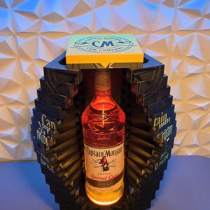 Könnte beinhalten: Eine Flasche Captain Morgan Spiced Gold Rum in einem dekorativen schwarzen und gelben Display. Die Flasche wird von unten beleuchtet, und das Display trägt die Aufschrift "Captain Morgan" und "Spiced Gold". Das Display hat auch den Text "CM Rum Company".