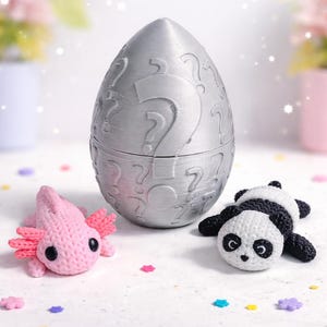 Può includere: Un contenitore argentato a forma di uovo con punti interrogativi, accanto a una statuina di axolotl rosa e una statuina di panda bianco e nero. L'axolotl ha branchie rosa e il panda è sdraiato.